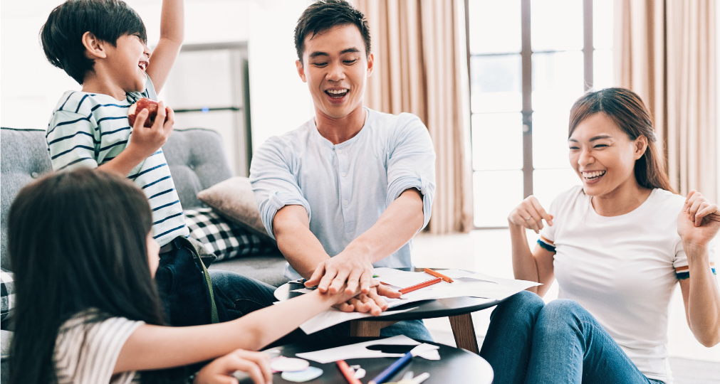 A family laughs and smiles while playing a game with creative materials.