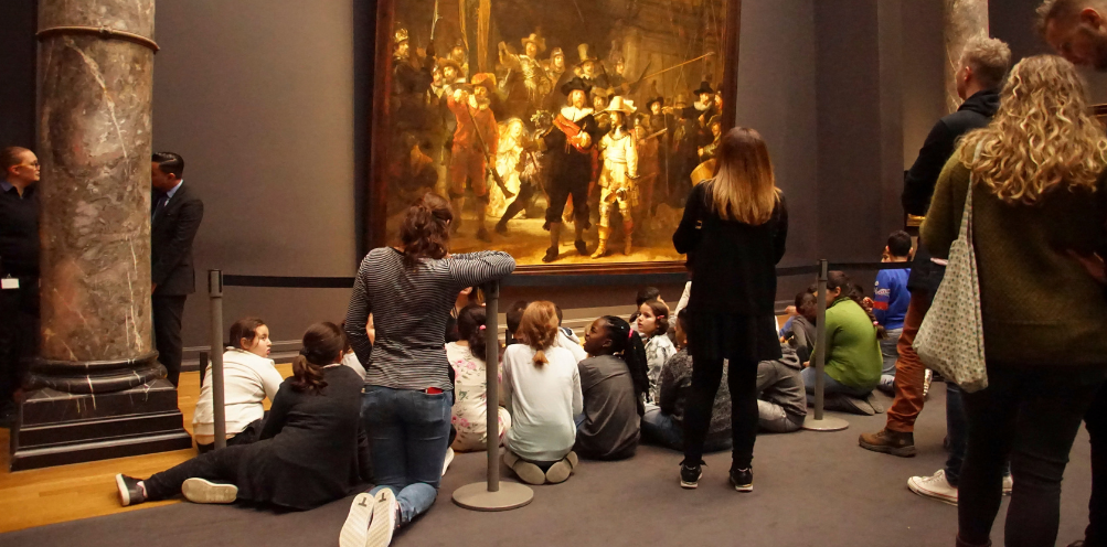 A group of students sits in front of Rembrandt's painting The Night Watch to study it.