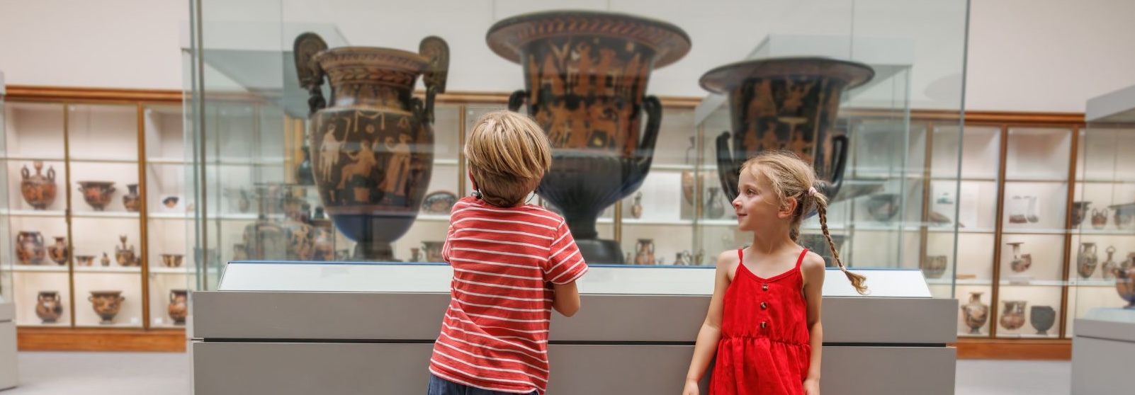 Two young children stand in front of a museum case holding ancient Greek vases.